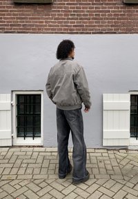 Szürke bomber dzseki bordázott mandzsettákkal, sötét farmernadrággal párosítva. A háttérben téglafal és fekete rácsos ablak látható.