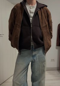 Giacca bomber in suede marrone sopra un hoodie nero, abbinata a jeans a gamba larga blu chiaro. Interno di una stanza con pareti bianche sullo sfondo.