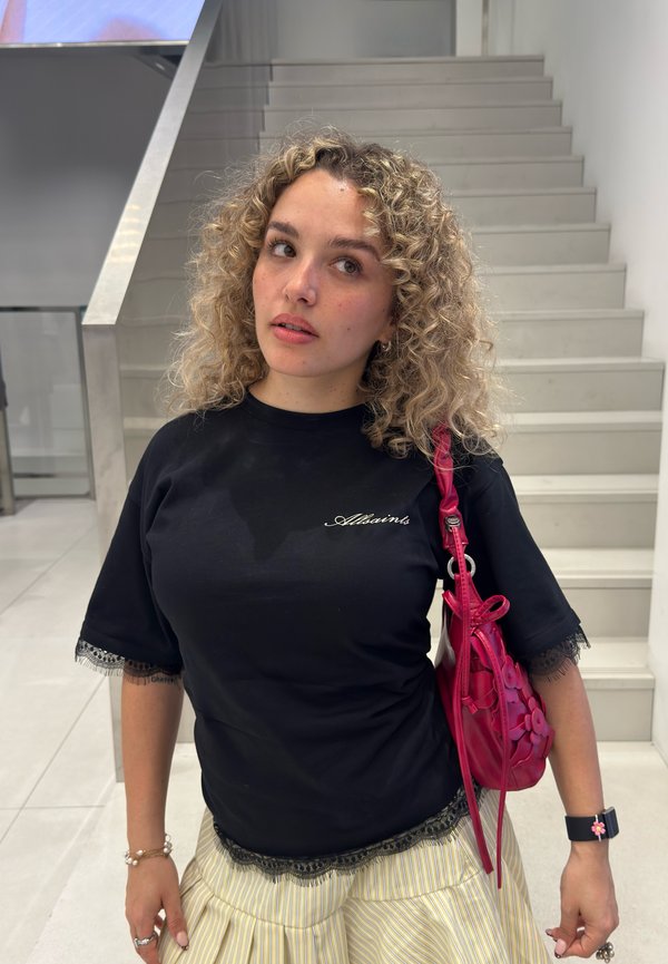 Mujer con cabello rubio rizado lleva una camiseta negra de AllSaints, falda a rayas, bolso rosa de hombro y un reloj inteligente negro, de pie junto a unas escaleras en el interior.