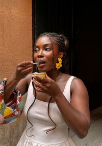 Donna con capelli intrecciati e orecchini a forma di fiore giallo che mangia da una piccola tazza verde utilizzando un cucchiaio bianco, in piedi vicino a un muro marrone.