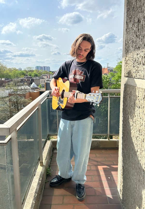 Guitarra acústica com um corpo de madeira clara e braço escuro, segurada por uma pessoa vestindo uma t-shirt preta, jeans claros, e sapatos pretos, numa varanda.