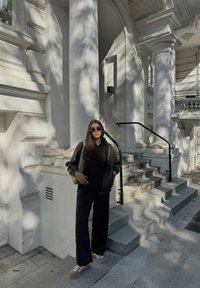 Μαύρο φερμουάρ μπουφάν πάνω από μια μαύρη φούτερ, μαύρο παντελόνι σε φαρδιά γραμμή και καφέ αθλητικά παπούτσια. Στέκεται σε πέτρινα σκαλοπάτια κοντά σε λευκούς αρχιτεκτονικούς κίονες.