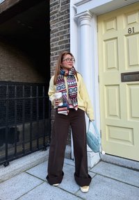 Femme portant une écharpe à motifs et un pull jaune, tenant une tasse de café et un sac bleu, debout près d'une porte jaune numérotée 81.