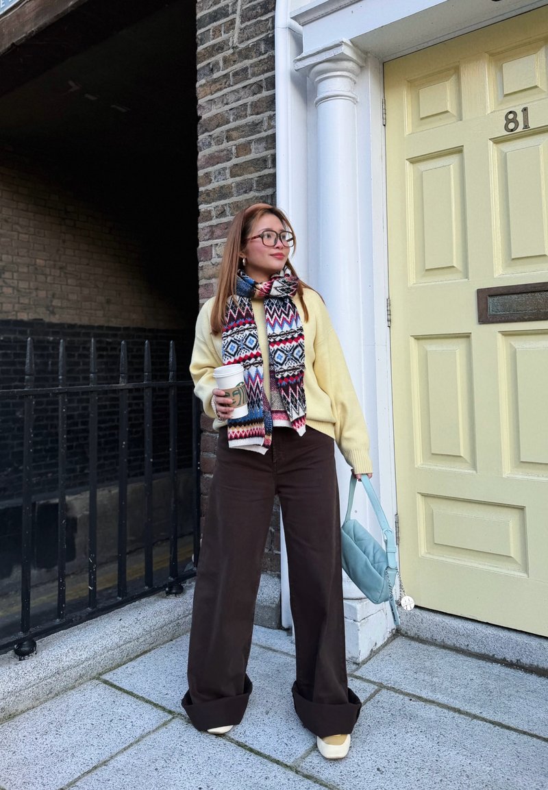 Γυναίκα με φουλάρι με σχέδια και κίτρινο πουλόβερ, κρατώντας ένα φλιτζάνι καφέ και μια μπλε τσάντα, στέκεται δίπλα σε μια κίτρινη πόρτα με αριθμό 81.