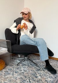 Sedia girevole in velluto a coste nero con base cromata, modello con camicia bianca a maniche lunghe, coprispalle grigio a maglia, jeans azzurri, che tiene un oggetto arancione.