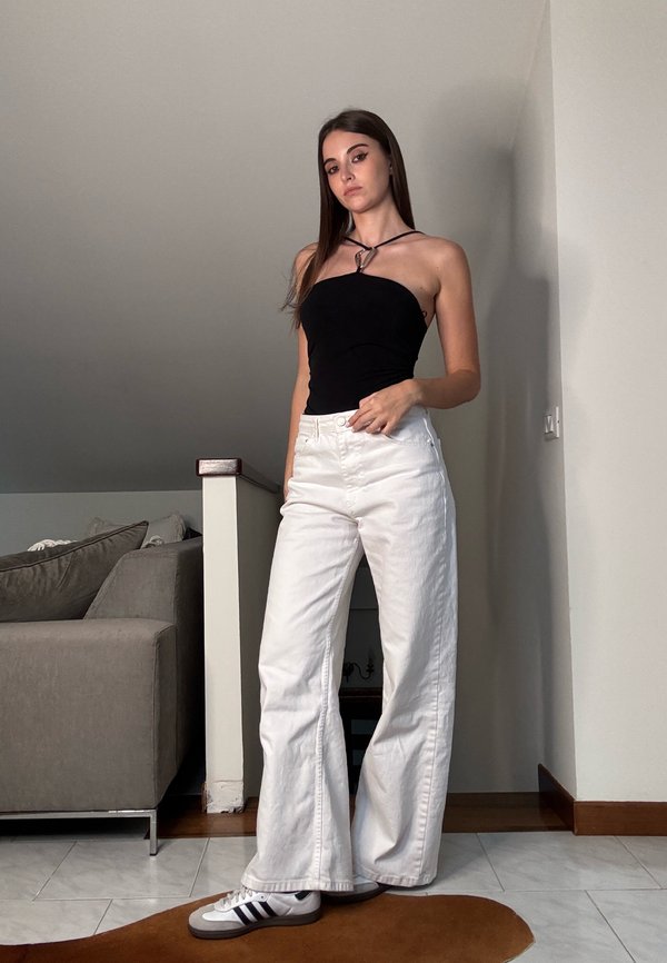 Black sleeveless top with white wide-leg jeans. The jeans feature a textured fabric. Adidas trainers with three black stripes, standing on a patterned rug.