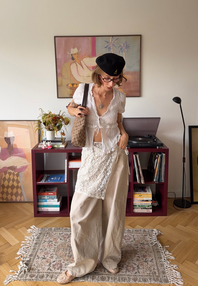 Donna con occhiali, berretto nero, top bianco in pizzo e pantaloni beige larghi, in piedi nel soggiorno con quadri, giradischi, libri e fiori.