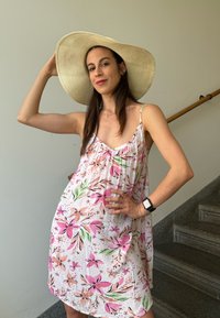 Φλοράλ φόρεμα με ροζ και πράσινα σχέδια, χωρίς μανίκια, ελαφρύ ύφασμα, σε συνδυασμό με ένα καπέλο από ψάθα με φαρδύ γείσο.