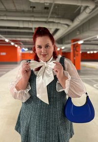 Pelēks teksturēts kleitas ar baltām plūstošām piedurknēm un lielu baltu loku pie apkakles, turot zilu rokassomu autostāvvietā.