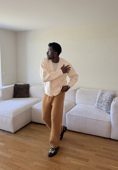 Hombre de pie en la sala de estar con un cárdigan beige, pantalones marrones y zapatos con estampado, ajustándose la manga mientras mira hacia un lado.
