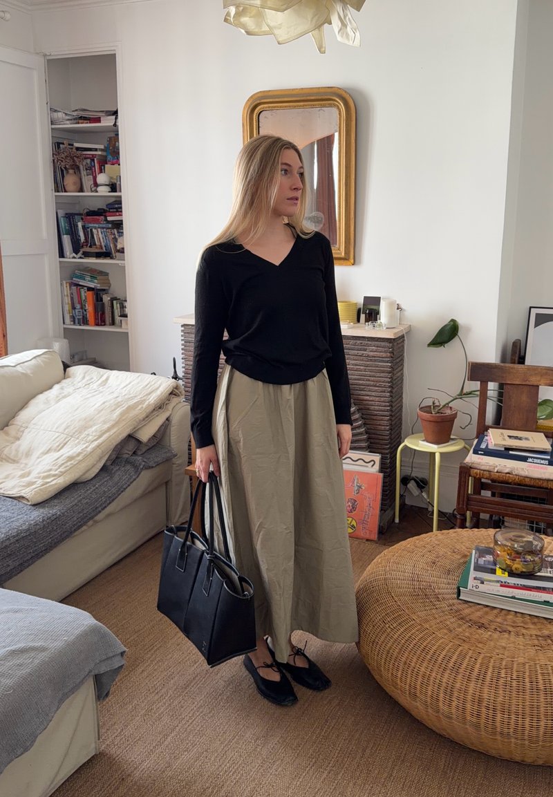 Femme en haut noir et jupe beige tenant un sac à main noir, debout dans un salon cosy avec des livres, des plantes et un ottoman en osier.