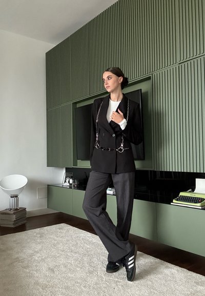 Blazer negro de doble botonadura con acento de cadena plateada, camisa blanca de manga larga, pantalones de pierna ancha grises y zapatillas negras con franjas blancas.