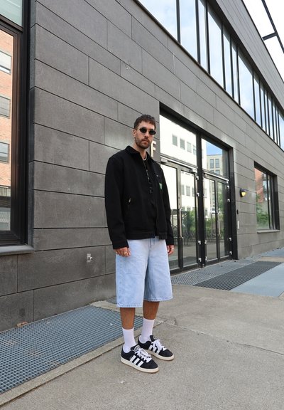 Chaqueta negra con cremallera, pantalones cortos de mezclilla claros, zapatillas negras con detalles blancos, calcetines blancos. De pie contra una pared gris, con puertas de cristal al fondo.