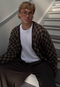 Cárdigan a cuadros marrón y negro sobre una camiseta blanca, combinado con pantalones oscuros de corte holgado, sentado en escaleras blancas. Usa gafas.