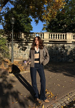 Chaqueta de diseño de pata de gallo, suéter a rayas, jeans oscuros de campana, zapatos marrones, sosteniendo una taza de café, contra un muro de piedra y hojas de otoño.