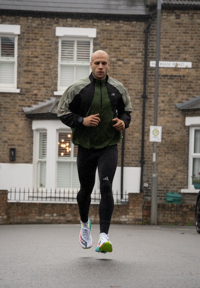 Läufer in einer schwarzen und grünen Jacke, schwarzen Leggings und bunten Laufschuhen. Nasser Asphalt und Ziegelmauerwerk mit Gebäuden im Hintergrund sichtbar.
