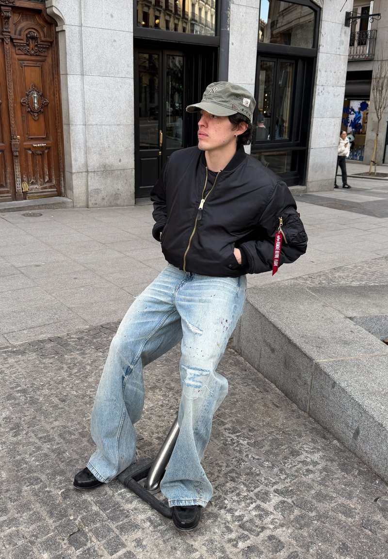 Junge Person in schwarzer Bomberjacke, weit geschnittenen hellblauen Jeans und grüner Mütze, die an einem Fahrradständer in einer Stadtstraße lehnt.
