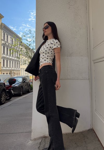 Mujer con gafas de sol negras, top blanco con lunares, vaqueros negros de cintura alta y botas, de pie en una acera de la ciudad frente a una pared blanca.
