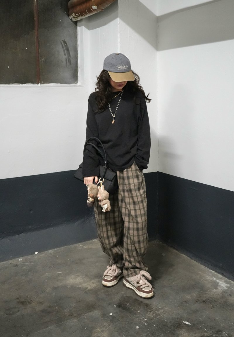 Personne portant une casquette grise et beige, un sweat-shirt noir, un pantalon à carreaux, des baskets en peluche, tenant un sac noir avec deux porte-clés en forme d'oursons.