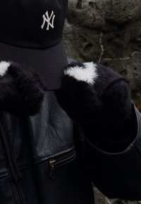 Șapcă de baseball neagră cu logo alb, mănuși negre din blană artificială cu accente albe și o jachetă neagră din piele matlasată cu buzunar cu fermoar.