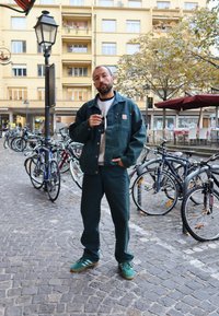 Zöld dzseki és hozzáillő nadrág erős anyagból. Az illető napszemüveget tart a kezében, és zöld sportcipőt visel. Kockakőből készült utca kerékpárokkal.
