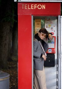 Κόκκινο θυρίδιο τηλεφώνου με χρυσή πινακίδα "ΤΗΛΕΦΩΝΟ", που διαθέτει τηλεφωνική μηχανή, αυτοκόλλητα και ένα άτομο με γκρι παλτό και γυαλιά ηλίου, που γέρνει μέσα.