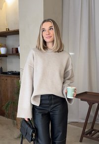 Γυναίκα με ξανθά μαλλιά που φοράει μπεζ πουλόβερ και μαύρο δερμάτινο παντελόνι, κρατώντας μια κούπα καφέ και μια μαύρη τσάντα σε ένα ζεστό εσωτερικό χώρο.