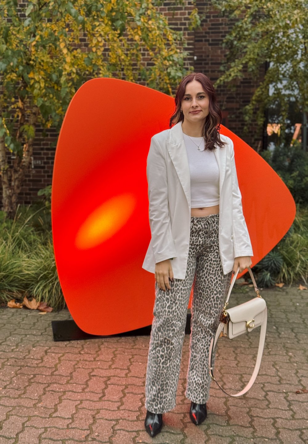 Bílý blazer, zkrácený bílý top, kalhoty s leopardím vzorem, černé podpatky. Drží krémovou kabelku. Za ní velká oranžová abstraktní socha.