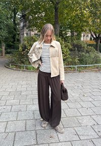 Giacca scamosciata di un beige chiaro con frange, maglietta a righe, pantaloni larghi marroni e scarpe con stampa animale. Tiene una borsa scura intrecciata.