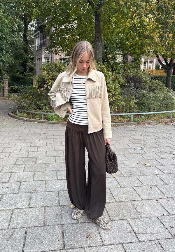 Ljusbeige mockajacka med fransar, randig topp, bruna vida byxor och skor med djurmönster. Håller en mörk, vävd handväska.