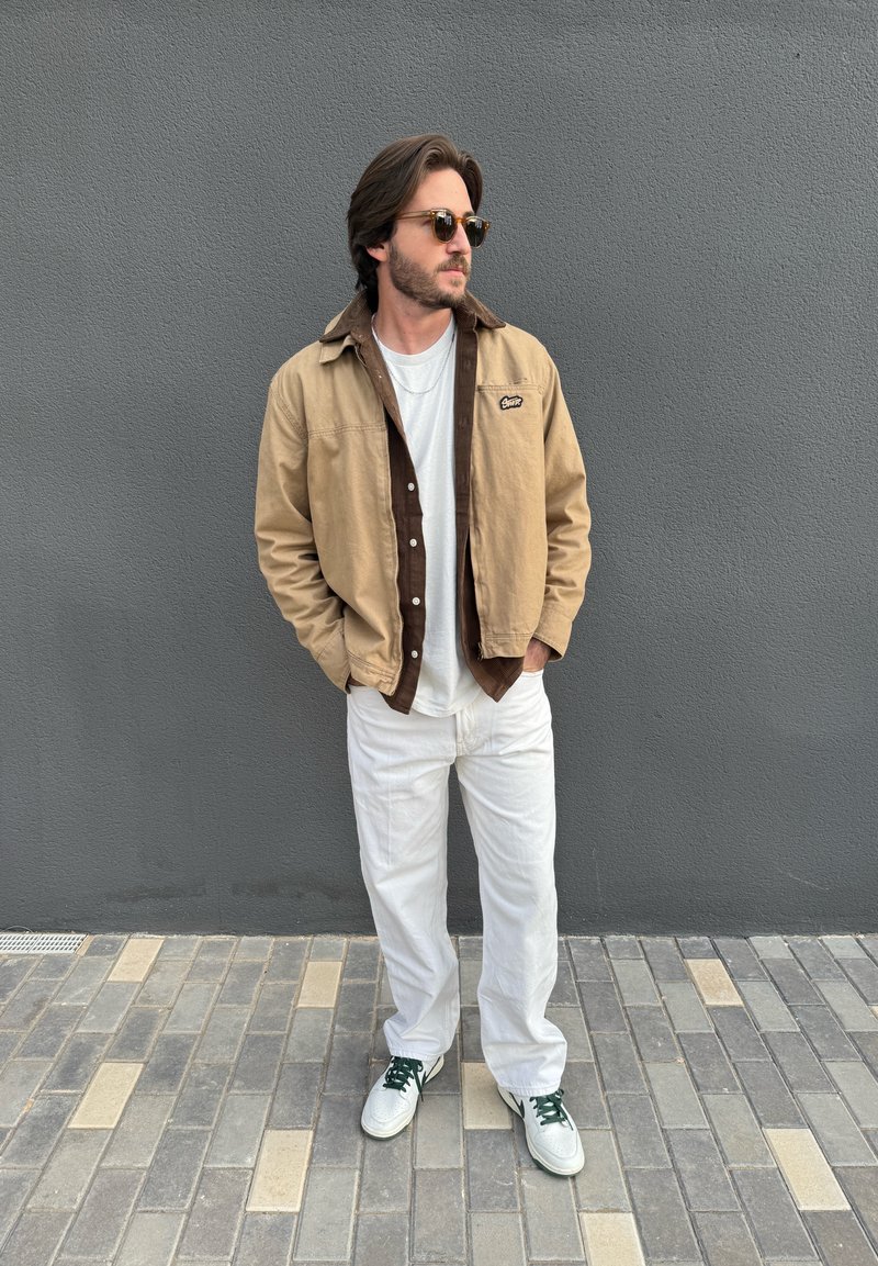 Tan jacket with a brown corduroy collar, white T-shirt, beige trousers, and white trainers with green accents. Background is a grey wall.