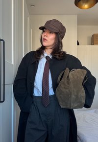 Casquette marron, manteau foncé, chemise blanche et cravate rayée. Pantalon gris taille haute et sac en suède vert. Fond intérieur simple.