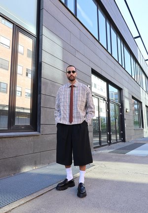 Camisa de cuadros con gran cuello, corbata marrón, pantalones cortos negros a rayas, calcetines blancos y zapatos negros robustos, situados frente a un edificio moderno.
