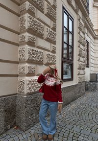 Rode gebreide trui met witte en blauwe patroon op het schouderstuk, gecombineerd met lichtblauwe spijkerbroek. Muren zijn voorzien van gestructureerde steen met decoratieve details.