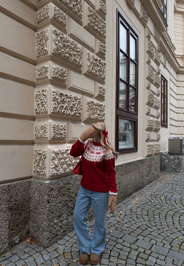 Rode gebreide trui met witte en blauwe patroon op het schouderstuk, gecombineerd met lichtblauwe spijkerbroek. Muren zijn voorzien van gestructureerde steen met decoratieve details.
