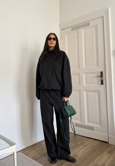 Mujer con chaqueta negra, pantalones y gafas de sol, sosteniendo un pequeño bolso verde, de pie cerca de una puerta blanca con paneles en el interior.