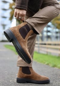 Persona uzvelk brūnos zamša Čelsija zābakus ar melnu zoli, ģērbjot béžus bikses un sudraba gredzenu ārā uz bruģēta ceļa.