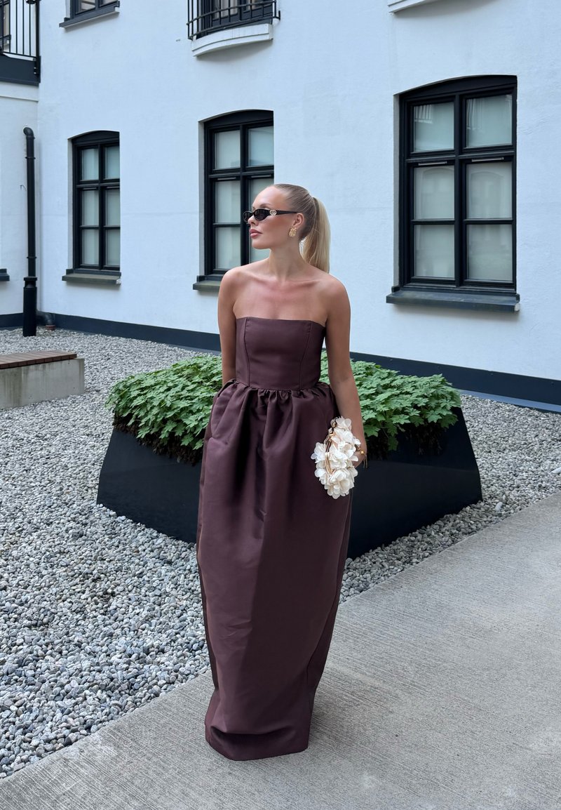 Femme avec queue de cheval blonde portant une robe marron foncé sans bretelles et des lunettes de soleil, tenant un bouquet de fleurs blanches, debout près d'une jardinière devant un bâtiment blanc.