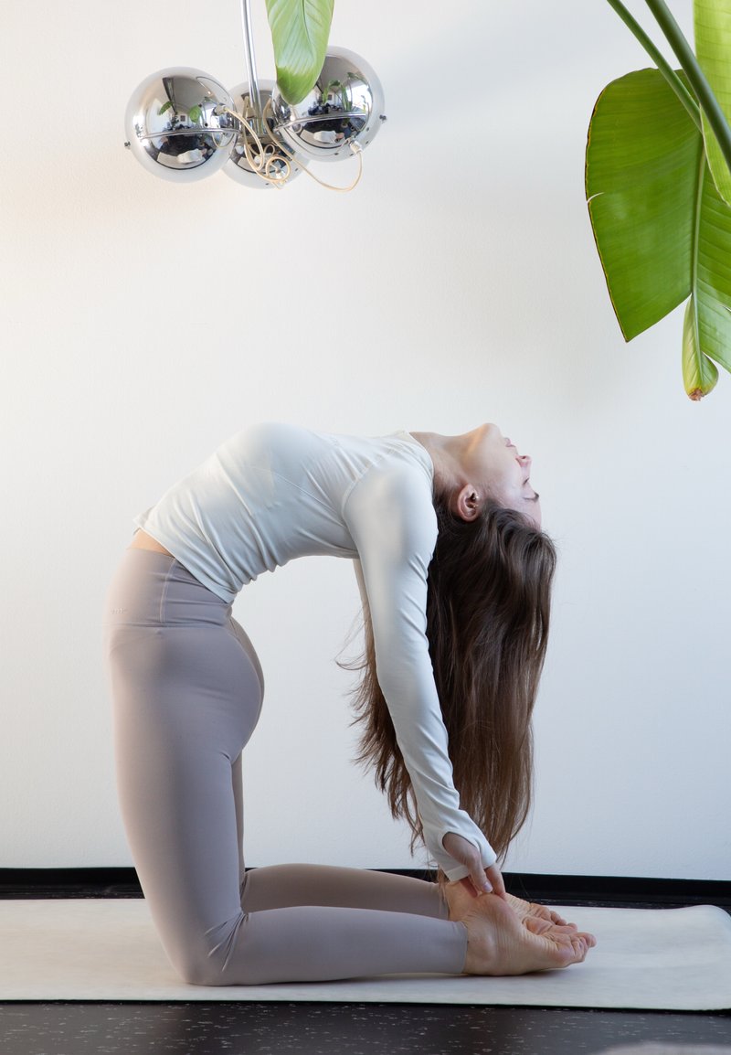 Femme en haut blanc à manches longues et legging beige réalisant la posture de yoga du chameau sur un tapis à l'intérieur, près de plantes et d'un luminaire argenté.