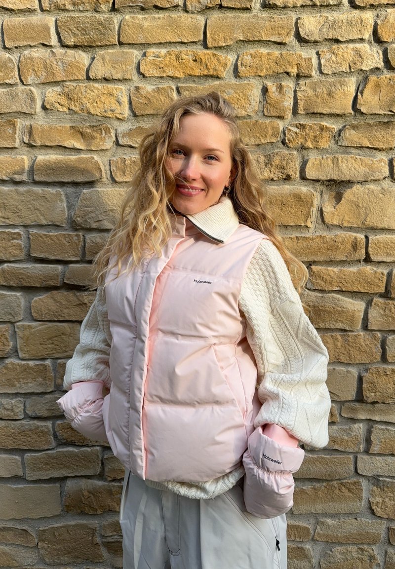 Gepolsterte rosa Weste mit hohem Kragen, Steppmuster und weißen, kabelgestrickten Ärmeln. Mit Logodetail auf der linken Seite.
