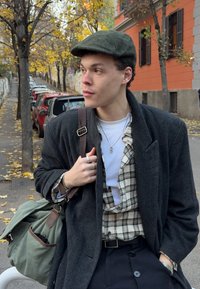 Junger Mann mit grauer Schiebermütze, dunklem Mantel, kariertem Hemd und einer grünen Tasche auf einer herbstlichen Straße mit gelben Blättern.