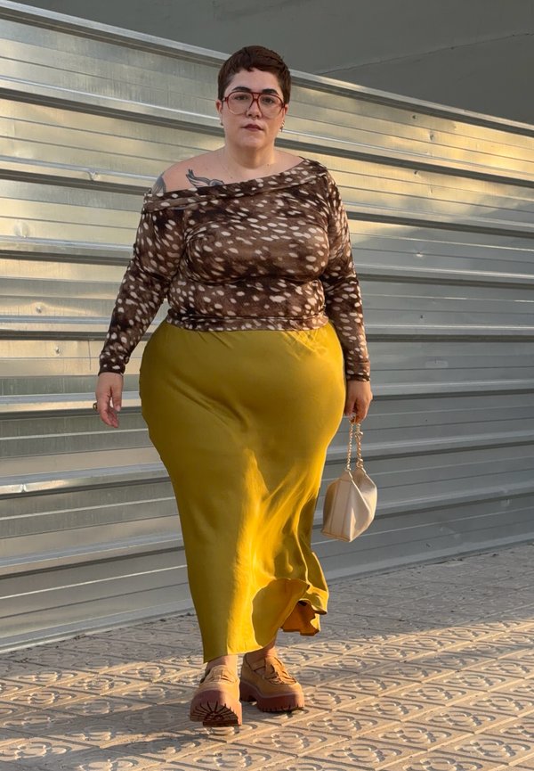 Femme aux cheveux courts et lunettes marchant à l'extérieur, portant un haut marron à pois, une jupe jaune et tenant un petit sac à main beige.