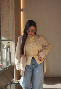 Beige blazer over en lys gul skjorte med knapper, parret med løse blå jeans. Tilbehøret inkluderer store klare briller og en mønstrete håndveske.