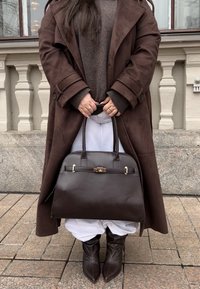 Bruine leren handtas met een ronde vorm, gouden beslag en twee handvatten. Draagt een lange bruine jas en witte broek. Gelaagde laarzen.