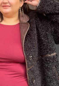 Musta teksturoitu takki, jossa on ruskeat yksityiskohdat ja napit, puettuna viininpunaisten yläosan päälle. Lähikuva henkilön olkapäästä ja korvakorusta on nähtävissä.