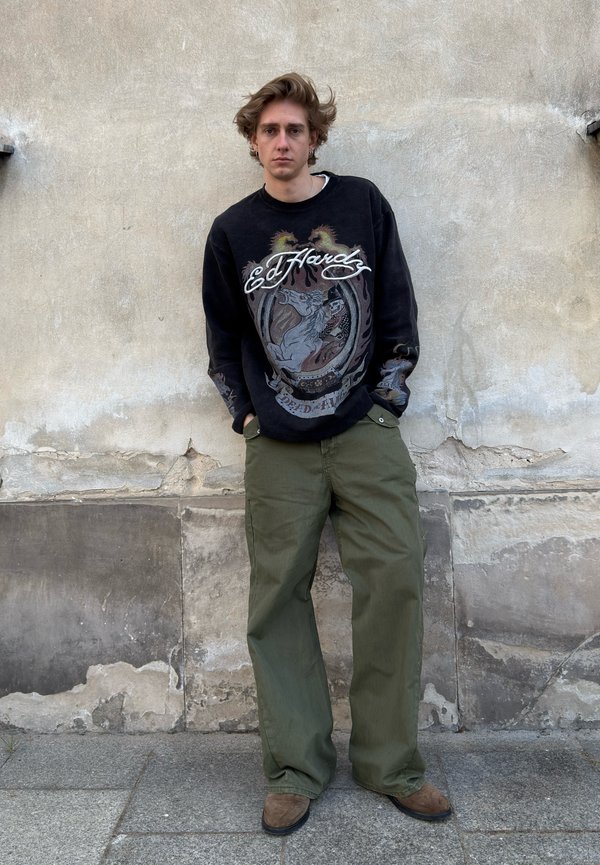 Jonge man met warrig haar die een zwart Ed Hardy sweatshirt, groene losse broek en bruine schoenen draagt, staat tegen een versleten betonnen muur.