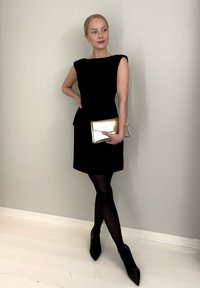 Vestido negro sin mangas con cintura peplum, combinado con un clutch dorado y medias negras translúcidas. Tacones negros puntiagudos completan el conjunto.
