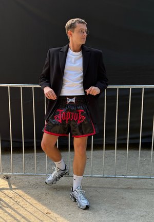 Persona vestida con una chaqueta negra sobre una camiseta blanca, pantalones cortos negros con acentos rojos y texto, y zapatillas deportivas plateadas con calcetines blancos.