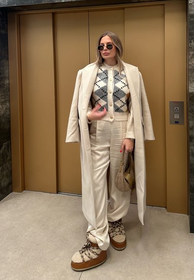 Abrigo largo color crema sobre un suéter estampado, pantalones crema y botas distintivas con parte superior texturizada. Los accesorios incluyen gafas de sol y un bolso.