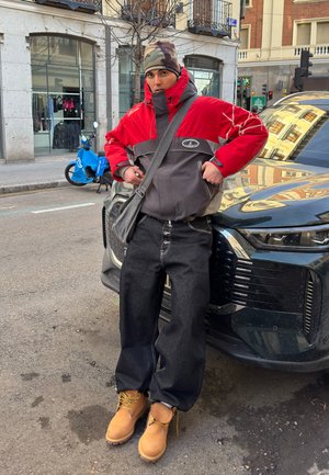 Hombre con chaqueta roja y gris, gorra de camuflaje, jeans negros anchos y botas color canela apoyado contra un coche negro en una calle urbana.